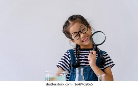 Childhood Girl Wearing Glasses Smiling And Holding Magnifying Glass While Doing Science Experiment At Table In Classroom Looking At Camera. Innocence Kid With Test Tube Front View. Education Concept.