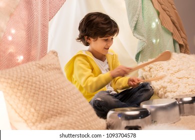 Childhood And Hygge Concept - Happy Little Boy With Cooking Pots Playing Music In Kids Tent At Home