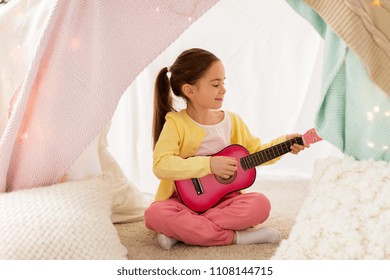 childhood and hygge concept - happy little girl playing toy guitar in kids tent at home