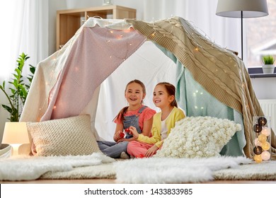 Childhood And Hygge Concept - Happy Little Girls Playing Tea Party With Toy Crockery In Kids Tent At Home