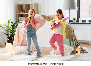 Childhood, Hygge And Friendship Concept - Happy Girls With Guitar And Microphone Playing Music And Singing Near Kids Tent Or Teepee At Home