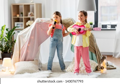 Childhood, Hygge And Friendship Concept - Happy Girls With Guitar And Microphone Playing Music And Singing Near Kids Tent Or Teepee At Home