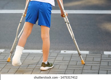 Childhood Injuries. Young Boy In Orthopedic Cast On Crutches Walking On The Street Near The Road. Child With Broken Leg On Crutches, Ankle Injury. Bone Fracture And Ankle Fracture 