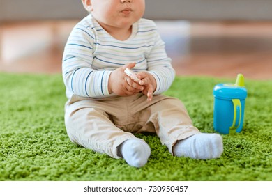 Childhood, Kids And People Concept - Baby Boy Sitting On Floor With Sippy Cup And Eating Rice Cracker At Home