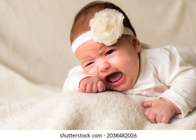 Childhood, Motherhood, Emotions, Fashion Concept - Worried Sad Crying Chubby Baby With Open Mouth Close Up Little Girl In Beautiful White Dress Floral Headband Lying On Soft Warm Bed On Tummy At Home