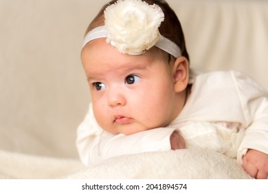 Childhood, Motherhood, Emotions, Fashion Concept - Worried Sad Crying Chubby Baby With Open Mouth Close Up Little Girl In Beautiful White Dress Floral Headband Lying On Soft Warm Bed On Tummy At Home