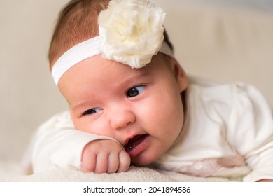 Childhood, Motherhood, Emotions, Fashion Concept - Worried Sad Crying Chubby Baby With Open Mouth Close Up Little Girl In Beautiful White Dress Floral Headband Lying On Soft Warm Bed On Tummy At Home