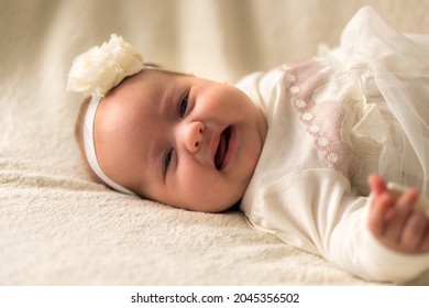 Childhood, Motherhood, Emotions, Fashion Concept - Worried Sad Crying Chubby Baby With Open Mouth Close Up Little Girl In Beautiful White Dress Floral Headband Lying On Soft Warm Bed On Tummy At Home