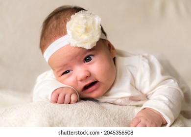 Childhood, Motherhood, Emotions, Fashion Concept - Worried Sad Crying Chubby Baby With Open Mouth Close Up Little Girl In Beautiful White Dress Floral Headband Lying On Soft Warm Bed On Tummy At Home