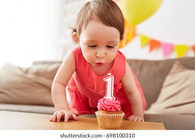 childhood, people and celebration concept - happy baby girl blowing to candle with number 1 on cupcake on birthday party at home