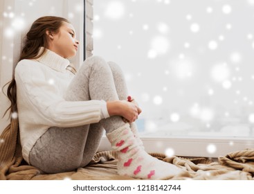 Childhood, Sadness And People Concept - Sad Beautiful Girl In Sweater Sitting On Sill At Home Window In Winter