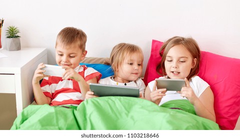 childhood, technology and family concept - little kids with tablet pc computer and smartphones in bed at home