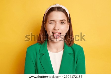 Childish behavior. Playful positive woman wearing green jacket winking to camera showing tongue out posing isolated over yellow background.