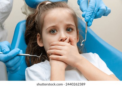 Childish fear. Cute little girl sitting in the dentist chair and covering her mouth in fear, being afraid of a dental drill in the hands of her doctor - Powered by Shutterstock