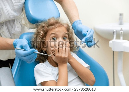 Childish fear. Cute little girl sitting in the dentist chair and covering her mouth in fear, being afraid of a dental drill in the hands of her doctor