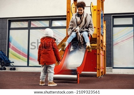 Childish young woman ride a slides at a child playground. A little boy looking at her.