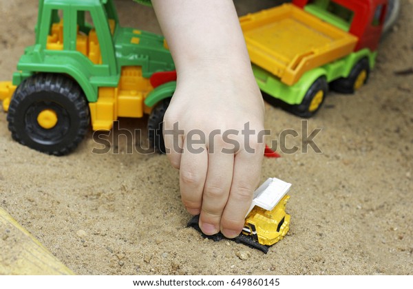 Children in air in the\
warm season. The hand of a child playing with plastic building toy\
cars in the sand.