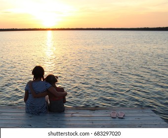 Children With Arms Around Each Other Sitting On The Dock At Sunset