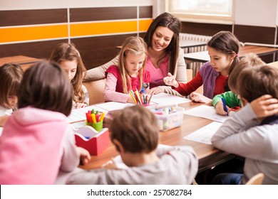 Children Boys And Girls Sitting Together Around The Table In Classroom And Drawing. With Them Is Their Young And Beautiful Teacher. 