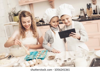 Children In Chef's Hats With Mother In Kitchen. Mother Is Putting Dough In Baking Dish And Kids Are Looking On Tablet.
