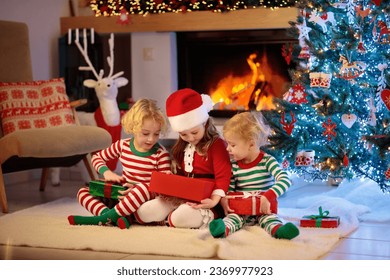 Children at Christmas tree and fireplace on Xmas eve. Family with kids celebrating Christmas at home. Boy and girl in matching pajamas decorating xmas tree and opening presents. Holiday gifts for kid. - Shutterstock ID 2369977923