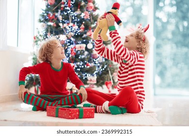 Children at Christmas tree and fireplace on Xmas eve. Family with kids celebrating Christmas at home. Boy and girl in matching pajamas decorating xmas tree and opening presents. Holiday gifts for kid. - Shutterstock ID 2369992315