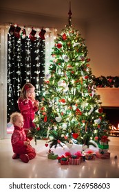 Children At Christmas Tree And Fireplace On Xmas Eve. Family With Kids Celebrating Christmas At Home. Boy And Girl In Matching Sweater Decorating Xmas Tree And Opening Presents. Holiday Gifts For Kid.