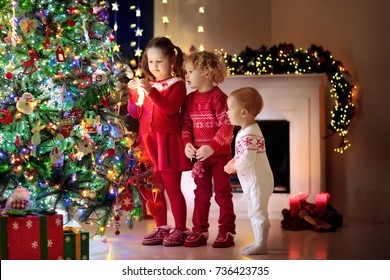 Children At Christmas Tree And Fireplace On Xmas Eve. Family With Kids Celebrating Christmas At Home. Boy And Girl In Matching Sweater Decorating Xmas Tree And Opening Presents. Holiday Gifts For Kid