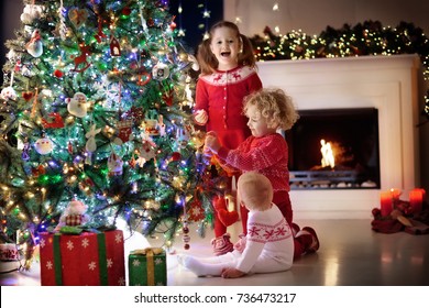 Children At Christmas Tree And Fireplace On Xmas Eve. Family With Kids Celebrating Christmas At Home. Boy And Girl In Matching Sweater Decorating Xmas Tree And Opening Presents. Holiday Gifts For Kid.