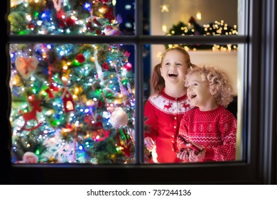 Children At Christmas Tree And Fireplace On Xmas Eve. Family With Kids Celebrating Christmas At Home. Boy And Girl In Matching Sweater Decorating Xmas Tree And Opening Presents. Holiday Gifts For Kid.