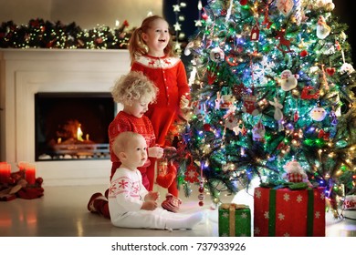 Children At Christmas Tree And Fireplace On Xmas Eve. Family With Kids Celebrating Christmas At Home. Boy And Girl In Matching Sweater Decorating Xmas Tree And Opening Presents. Holiday Gifts For Kid.