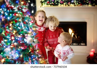 Children At Christmas Tree And Fireplace On Xmas Eve. Family With Kids Celebrating Christmas At Home. Boy And Girl In Matching Sweater Decorating Xmas Tree And Opening Presents. Holiday Gifts For Kid.