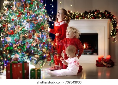 Children At Christmas Tree And Fireplace On Xmas Eve. Family With Kids Celebrating Christmas At Home. Boy And Girl In Matching Sweater Decorating Xmas Tree And Opening Presents. Holiday Gifts For Kid.