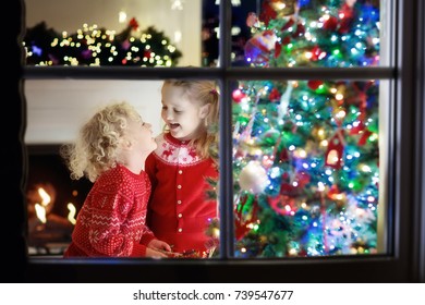 Children At Christmas Tree And Fireplace On Xmas Eve. Family With Kids Celebrating Christmas At Home. Boy And Girl In Matching Sweater Decorating Xmas Tree And Opening Presents. Holiday Gifts For Kid.