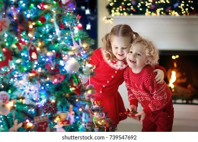 Children At Christmas Tree And Fireplace On Xmas Eve. Family With Kids Celebrating Christmas At Home. Boy And Girl In Matching Sweater Decorating Xmas Tree And Opening Presents. Holiday Gifts For Kid.