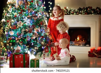 Children At Christmas Tree And Fireplace On Xmas Eve. Family With Kids Celebrating Christmas At Home. Boy And Girl In Matching Sweater Decorating Xmas Tree And Opening Presents. Holiday Gifts For Kid.