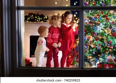 Children At Christmas Tree And Fireplace On Xmas Eve. Family With Kids Celebrating Christmas At Home. Boy And Girl In Matching Sweater Decorating Xmas Tree And Opening Presents. Holiday Gifts For Kid.