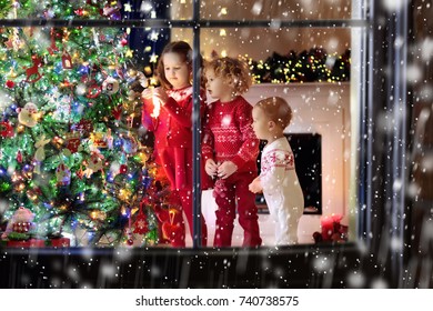 Children At Christmas Tree And Fireplace On Xmas Eve. Family With Kids Celebrating Christmas At Home. Boy And Girl In Matching Sweater Decorating Xmas Tree And Opening Presents. Holiday Gifts For Kid.