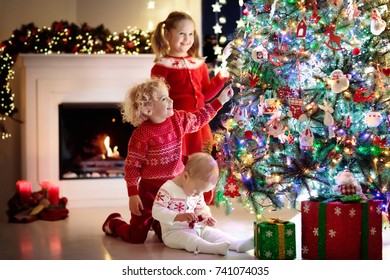 Children At Christmas Tree And Fireplace On Xmas Eve. Family With Kids Celebrating Christmas At Home. Boy And Girl In Matching Sweater Decorating Xmas Tree And Opening Presents. Holiday Gifts For Kid.