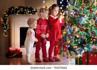 Children At Christmas Tree And Fireplace On Xmas Eve. Family With Kids Celebrating Christmas At Home. Boy And Girl In Matching Sweater Decorating Xmas Tree And Opening Presents. Holiday Gifts For Kid.