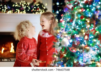 Children At Christmas Tree And Fireplace On Xmas Eve. Family With Kids Celebrating Christmas At Home. Boy And Girl In Matching Sweater Decorating Xmas Tree And Opening Presents. Holiday Gifts For Kid.