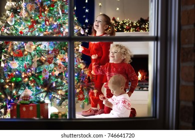 Children At Christmas Tree And Fireplace On Xmas Eve. Family With Kids Celebrating Christmas At Home. Boy And Girl In Matching Sweater Decorating Xmas Tree And Opening Presents. Holiday Gifts For Kid.