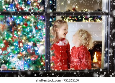 Children At Christmas Tree And Fireplace On Xmas Eve. Family With Kids Celebrating Christmas At Home. Boy And Girl In Matching Sweater Decorating Xmas Tree And Opening Presents. Holiday Gifts For Kid