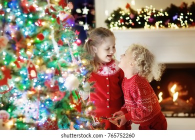 Children At Christmas Tree And Fireplace On Xmas Eve. Family With Kids Celebrating Christmas At Home. Boy And Girl In Matching Sweater Decorating Xmas Tree And Opening Presents. Holiday Gifts For Kid.