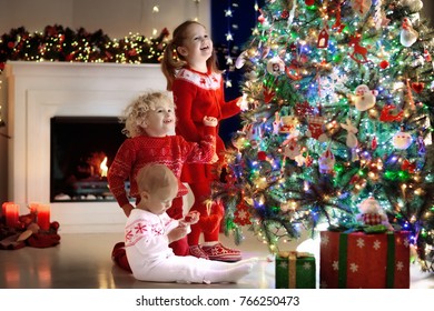 Children At Christmas Tree And Fireplace On Xmas Eve. Family With Kids Celebrating Christmas At Home. Boy And Girl In Matching Sweater Decorating Xmas Tree And Opening Presents. Holiday Gifts For Kid.