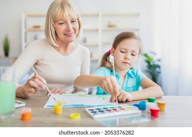 Children Creativity Concept. Portrait Of Little Girl Painting With Watercolors And Gouache At Home Together With Granny. Happe Mature Lady Helping Grandchild Creating Picture, Sitting At Table