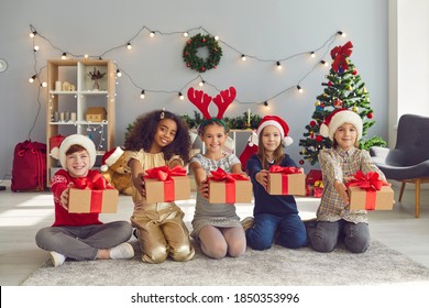 Children of different nationalities sit in a row and hold out Christmas presents in front of them in the living room with Christmas decorations. Concept of exchanging gifts, kindness and charity.
