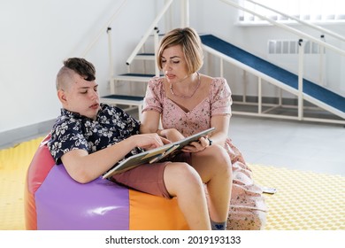 Children With Disability Reading Book And Communicating. Cerebral Palsy Boy Talking With Woman Tale Therapist. Rehabilitation Center, Mother With Teenage Person Learning To Speak, Pronounce At Home