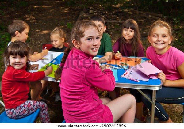 Children draw in nature. Lots of\
colored papers and pencils on a blue table. Boys and girls play\
outdoors. Drawing lesson in the park. Rest in a tent camp in the\
summer.