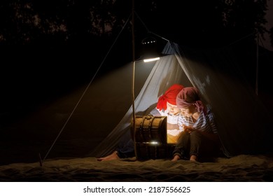 Children Dressed As Sailors Play In A Tent On The Sand At Night By The Light Of A Lantern And Pretend To Be Pirates Looking For Treasure On A Map. Funny Kids Dream Of Adventures And Travels.